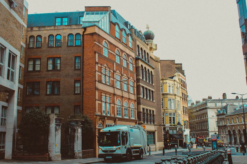 A street scene in an urban area featuring a mix of historic and modern buildings with brick facades, large windows, and ornate architectural details. In the foreground, a light blue waste collection vehicle from Waste Clearance Hammersmith is parked alongside the curb, ready for rubbish removal or private waste collection services. The vehicle's compact design and clean exterior indicate a professional waste management operation. Surrounding the vehicle, a row of black metal bike racks is aligned along the pavement, and nearby, black bollards provide separation between pedestrian and vehicle zones. To the left, there is an entrance with a gate surrounded by stone pillars, partially obscured by shrubbery, suggesting a private property or building access point. The sky is overcast, casting diffuse light over the scene, highlighting the textures of the brick and stone surfaces. Overall, the image depicts a typical city street used for urban rubbish collection, relevant to independent and alternative waste handling services like those offered by Waste Clearance Hammersmith, situated in a setting characteristic of central London or similar UK locations.
