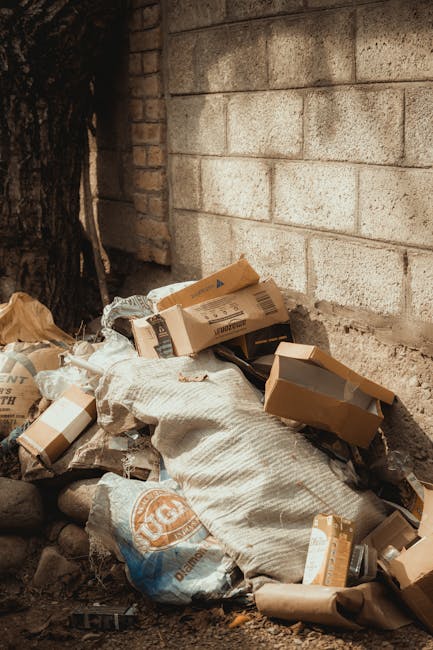 A pile of discarded cardboard boxes, some flattened and others partially intact, along with a large woven sack, all resting against a weathered brick wall on a rough, uneven ground surface with scattered small stones and dirt. The boxes are of varying sizes, mostly brown with printed labels, some slightly torn or crumpled, indicating recent or ongoing rubbish clearance. The woven sack is off-white with a textured surface and appears filled or partially filled. The background features a grey concrete block wall and a section of tree trunk with dark, textured bark visible to the left. The scene is outdoors, illuminated by natural light, suggesting an area used for waste disposal or private rubbish collection, consistent with independent trash clearance or alternative waste handling services like those offered by Waste Clearance Hammersmith.