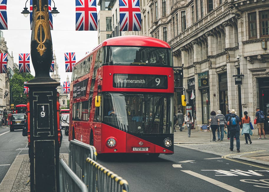 A red double-decker bus with the destination sign reading 'Hammersmith' and route number 9 is traveling along a city street lined with historic multi-story buildings featuring decorative stone facades and large windows. The bus's glossy surface reflects the surrounding environment, and its front windshield provides a clear view of the driver inside. On the left side, a black lamp post decorated with Union Jack flags extends upward, and in the foreground, a metal barrier separates the bus lane from the pedestrian walkway. Several pedestrians are visible on the sidewalk, some engaged in conversation, others walking or standing, dressed in casual attire suitable for mild weather. The scene is illuminated by natural daylight, casting soft shadows across the street. The overall setting suggests a busy urban area with ongoing public transportation activity, highlighting the importance of efficient rubbish removal services like those provided by Waste Clearance Hammersmith in maintaining cleanliness amid city traffic and pedestrian flow, especially in a historic and bustling central London environment.