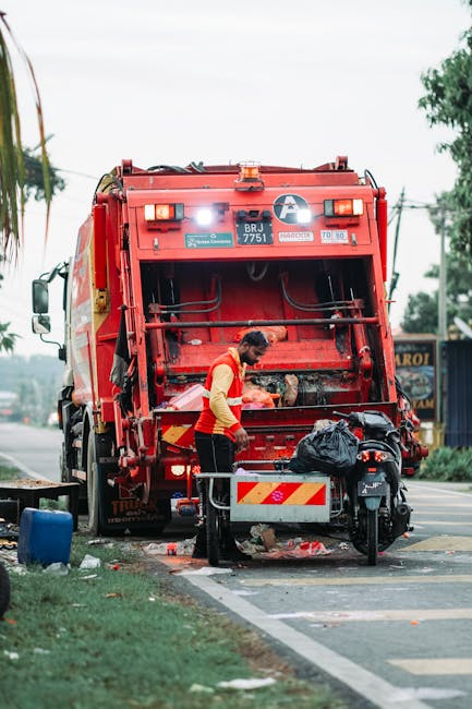 A waste collection worker wearing a yellow safety vest and black trousers is standing behind a red refuse collection vehicle, which is positioned on the side of a road. The vehicle's rear hatch is open, revealing internal mechanical parts and a large black trash bag resting on a metal bar. The worker appears to be sorting or clearing waste materials on the ground near the vehicle, with some debris visible around the area. The scene is set outdoors during daylight, with a partly cloudy sky, and in the background, there are trees, utility poles, and a small blue building or structure. The pavement is marked with white lines indicating parking or loading zones, and a bicycle is parked next to the waste bin on the curb. The image reflects a typical on-site rubbish collection operation, highlighting the practical aspects of private waste disposal services provided by companies like Waste Clearance Hammersmith.