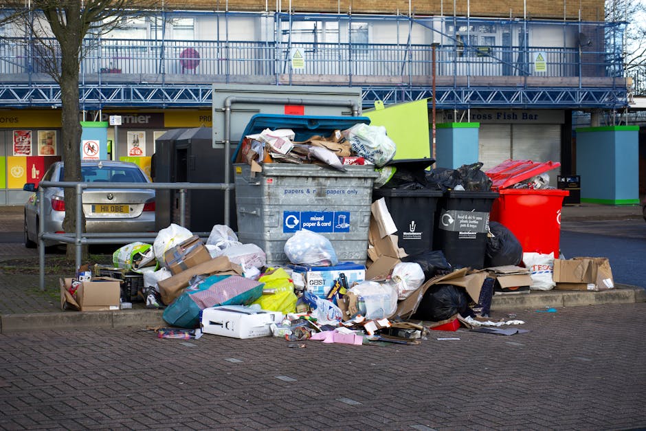 A pile of overflowing rubbish on a paved area in front of a commercial building, illuminated by daylight. The waste includes various black and red black bin bags, cardboard boxes, and paper-recycling containers. One large metal mixed paper and card waste bin is prominently situated in the center with an open lid, revealing crumpled paper, cardboard, and assorted packaging spilling out. Surrounding the bin are scattered cardboard boxes, plastic bags, and loose refuse including packaging materials and paper sheets, some of which are partially crushed or flattened. To the left, a parked silver car is visible, behind a metal railing that separates the waste area from the adjacent sidewalk. In the background, a building with large windows and a blue metal scaffold or balcony framework is visible alongside a storefront with signage and a closed shutter. The overall scene exemplifies a typical site of private waste accumulation awaiting rubbish removal or alternative waste collection services, as managed by Waste Clearance Hammersmith.