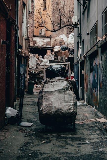 A narrow urban alleyway filled with a large accumulation of rubbish and debris. In the foreground, a large, wheeled rubbish bin with a weathered, light-colored fabric cover is positioned on the uneven, dark asphalt ground. Behind the bin, a clutter of cardboard boxes, plastic bags, and miscellaneous discarded items are stacked haphazardly against a back wall made of brick and concrete, partially obscured by leafless tree branches that extend overhead. The alley is flanked by two buildings: on the left, a reddish-brown brick wall with rusty metal features, and on the right, a grey concrete wall with a window secured by metal bars. Graffiti is visible on part of the right-side wall. The scene appears to be in a neglected area, with ambient lighting suggesting overcast skies or late afternoon. The image reflects a typical setting where private waste clearance, such as that provided by Waste Clearance Hammersmith, might be required to manage rubbish accumulation in urban backstreets, supporting the broader context of alternative waste handling methods for flats and shops in the city.