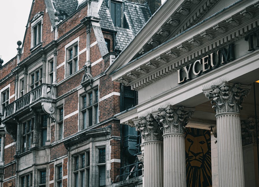 A close-up view of the façade of the Lyric Theatre, featuring classical architectural elements including four fluted Corinthian columns with ornate capitals supporting a decorative entablature. The sign above the entrance reads 'LYRIC' in bold black letters on a white background. To the left, there is an adjacent historic red brick building with multiple windows, some with decorative white stone surrounds and small metal balconies. The building's roofline displays slate tiles and ornamental details, and black raingutters run down the side. The environment is shaded, with diffused lighting highlighting the textures of the brickwork and stonework. The scene captures a section of urban architecture, suggesting a setting where external building debris is managed separately from the historic façade, relevant to private waste handling or alternative rubbish removal options that Waste Clearance Hammersmith might provide for such properties.