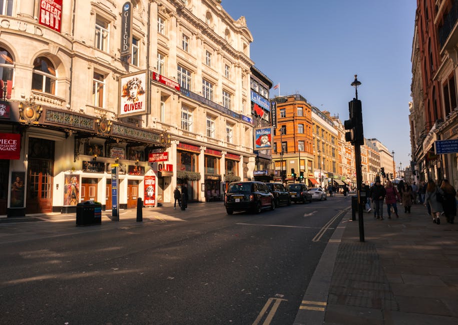 A busy urban street scene during daytime featuring a row of historic commercial buildings with ornate facades and signage, including a prominent theatrical poster advertising 'Oliver!'. The street has vehicles including black taxis and private cars parked or moving along the road, with pedestrians walking on the wide pavement. The background shows a clear blue sky and additional buildings with mixed architectural styles, some with red brick and others with cream or beige stone finishes. Street lamps are visible, and a traffic light is positioned at an intersection. The scene captures a lively city atmosphere with natural sunlight casting shadows, reflecting the typical setting for city centre rubbish removal and alternative waste handling services. Waste Clearance Hammersmith may occasionally operate in such urban environments to facilitate private rubbish clearance, especially around theatres or commercial zones, ensuring efficient and independent waste disposal outside local authority collection routes.