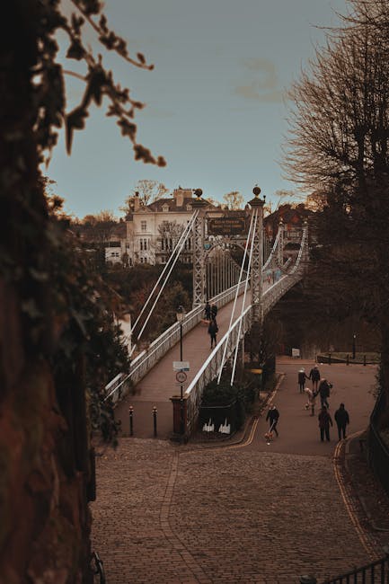 A view of a pedestrian suspension bridge spanning a river, with the entrance situated at a higher elevation on a cobbled slope. The bridge features white steel cables and railings, with a few individuals walking across it, some carrying bags or backpacks. On the riverbank below, there is a paved walkway with several people strolling, dressed in casual attire suitable for cool weather. Surrounding trees without leaves suggest late autumn or winter, and in the background, residential or historic buildings with light-colored facades and tiled roofs are visible against a pale sky during daytime. The scene appears to be part of an urban riverside area, where private or independent rubbish clearance services, such as those provided by Waste Clearance Hammersmith, might operate to support efficient waste management in the vicinity of such public infrastructure.