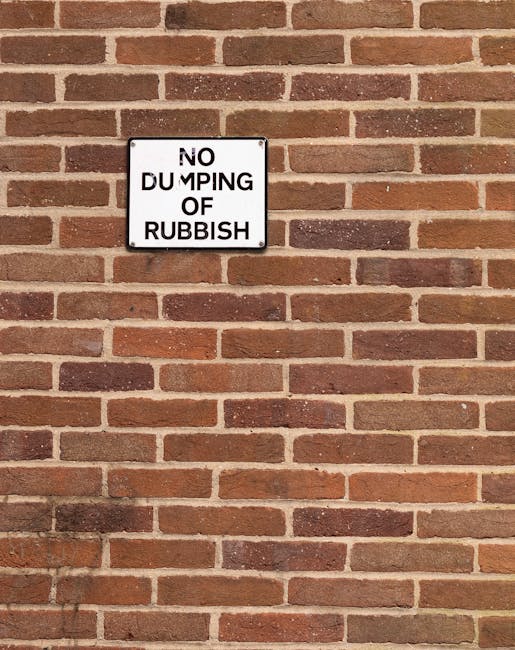 A rectangular white sign with black lettering reading 'NO DUMPING OF RUBBISH' is mounted on a red brick wall. The bricks vary in shades of reddish-brown and are arranged in a horizontal pattern with visible mortar joints. The sign is positioned towards the upper left section of the wall and is clearly visible against the textured brick background. The wall appears to be part of an outdoor setting, possibly a boundary wall or exterior surface associated with a property or waste management site. The natural lighting highlights the textures and colors of the bricks and the sign’s smooth surface, reflecting a neutral, professional aesthetic related to waste regulation and private waste handling practices.
