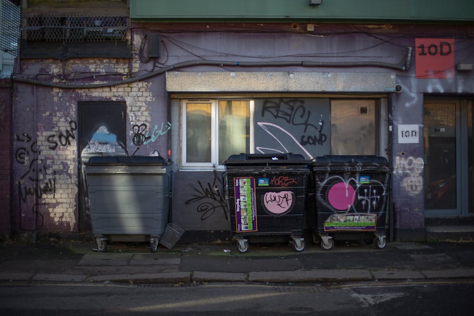 The image depicts an urban exterior wall with graffiti and weathered brickwork, situated behind a row of three large wheeled rubbish bins placed on the pavement. The left bin is made of grey plastic with a smooth surface, whereas the two on the right are black with colorful stickers, graffiti tags, and pink heart symbols on their surfaces. The bins are positioned directly beneath a window with a white frame, partially covered by a rolling shutter that is in a closed position. Above the window, there are visible electrical wires and conduit piping attached to the building's facade, which appears aged with peeling paint and marked by graffiti. To the right of the bins, there is a glass door with a visible red warning label, and adjacent to it, more graffiti and signs are painted or attached to the wall. The overall scene captures a typical backstreet setting, aligning with private waste collection or on-site clearance activities overseen by waste management services like Waste Clearance Hammersmith, amidst urban rubbish management context.